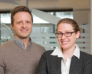A man and a woman smiling in front of a glass conference room window