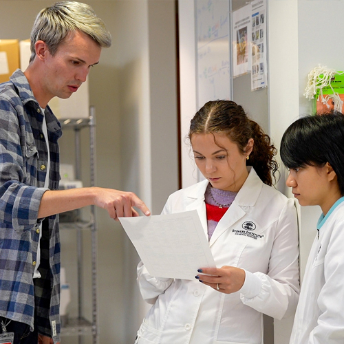 Man points finger at paper being held by a scientist in lab coat, and another scientist behind her