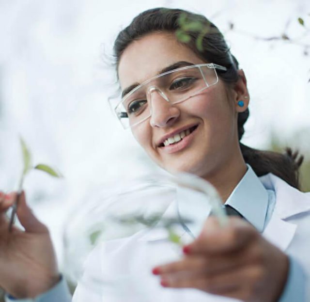 A closeup of a scientist smiling in safety glasses while inspecting a leaf