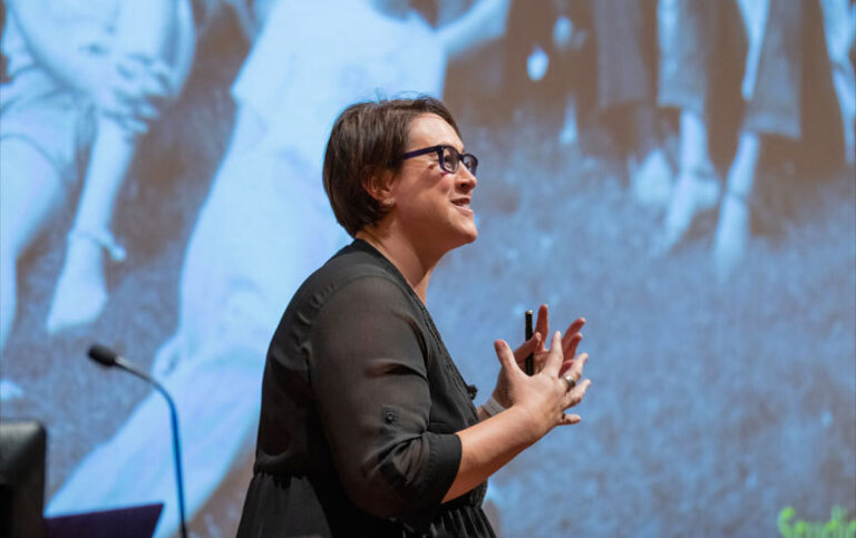 A woman wearing glasses on stage giving a lecture