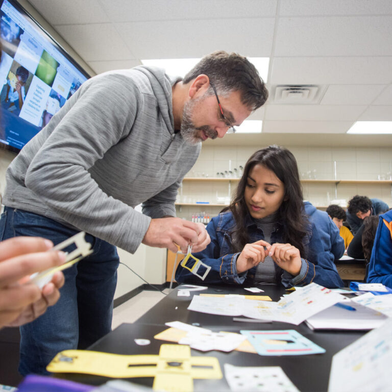 Man standing leaning over the student, seated at desk, explaining paper activity